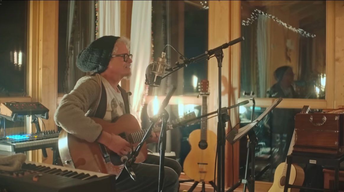 Niccolò Fabi con berretto di lana e occhiali mentre canta al microfono e suona la chitarra acustica nel suo studio di registrazione