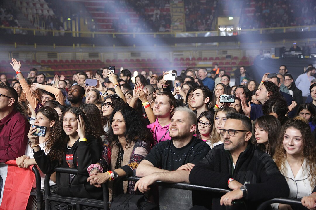 Folla numerosa al concerto di EuroFesta 2025 al chiuso, con persone che filmano il palco con gli smartphone vicino alla barriera.