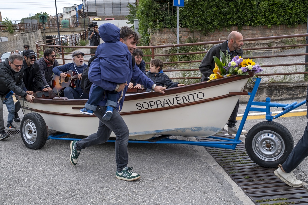Performers che spingono in strada una barca su ruote decorata con fiori con la scritta SOPRAVÈNTO durante il festival Sopravènto 2025, mentre un musicista suona la chitarra all'interno. Foto di Chiara Broccoli.