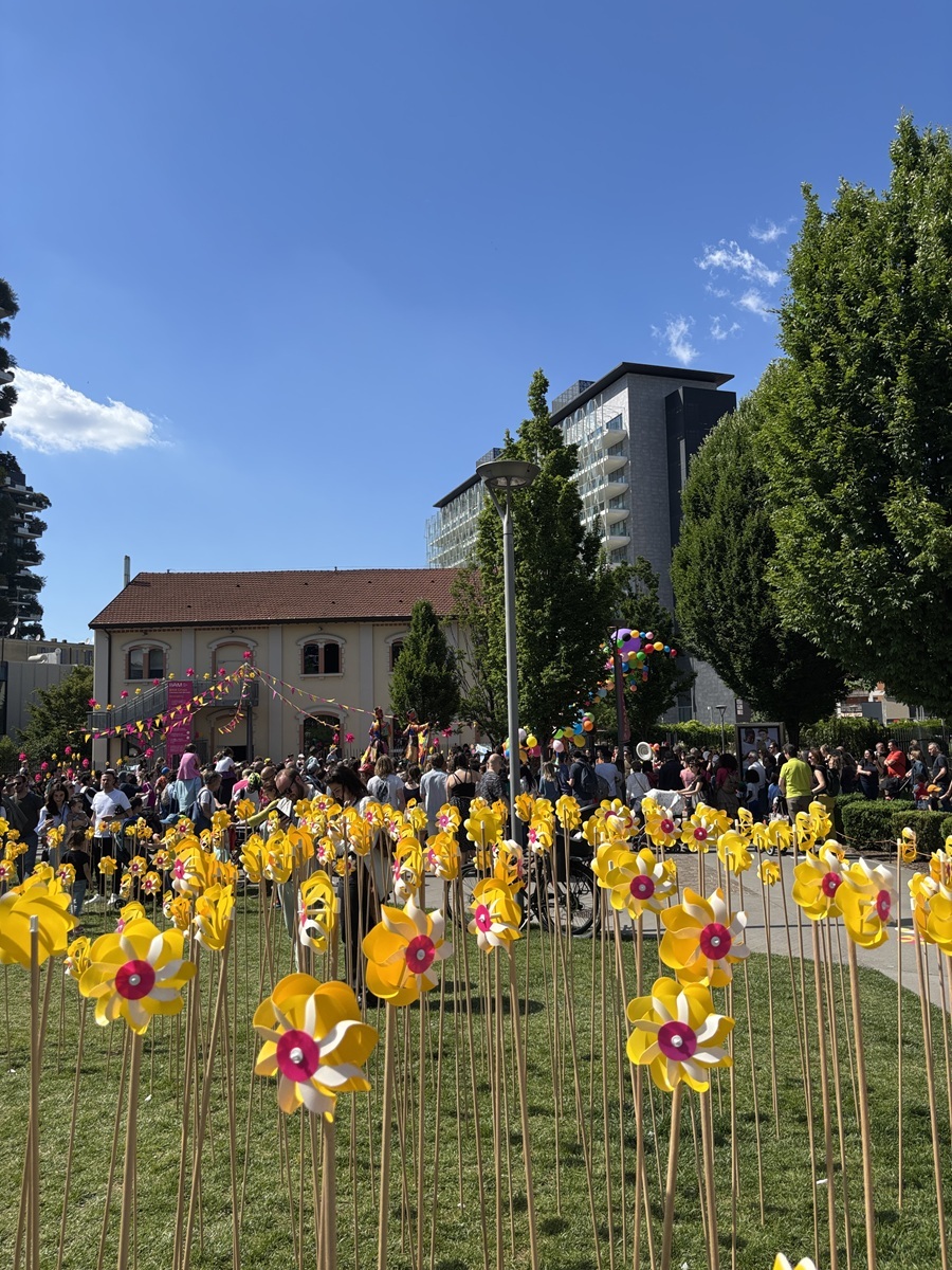 BAM biblioteca degli alberi. Girandole Fotografia scattata a livello del suolo a Milano, con una fitta distesa di girandole gialle e rosa su bastoncini in primo piano su un prato verde. Sullo sfondo, una folla partecipa a un evento all'aperto davanti alla Casa della Memoria (edificio giallo con bandierine) e all'edificio moderno BAM. Il cielo è blu e la scena è luminosa e festosa. foto realizzata da Yolande Duminuco